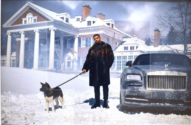 A winter scene, Drake, holding the leash of a dog, outside, standing beside a rolls royce car that has been out in the snow, and standing in front of a large house with white columns in the front 