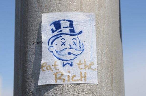 a small paste up on a utililty pole, blue stencil of the monopoly guy's head, top hat and X's for eyes, under his head are the words eat the rich
