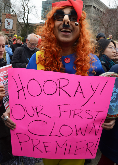 a man in an orange wig and clown costume holds a sign on pink bristol board that says Hooray! Our first clown premier 