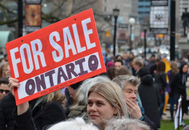 at a Queens Park demonstration on May day, a woman holds a red and white for sale sign with the word Ontario written in the blank, for sale Ontario