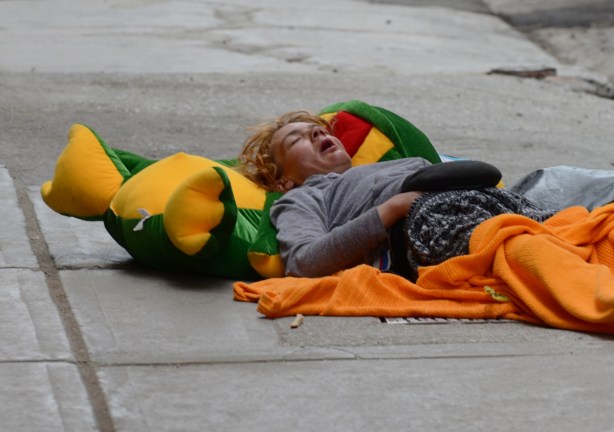 a person is sleeping on their back on the sidewalk. Their head is on a large green frog pillow 