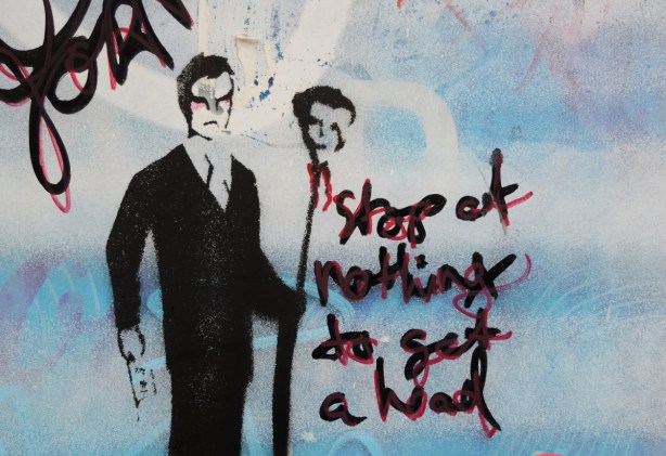 stencil on a wall that has been spray painted a pale blue, black ink, man holding a tall stick on the top of which is a head, words that say, stop at nothing to get a head 