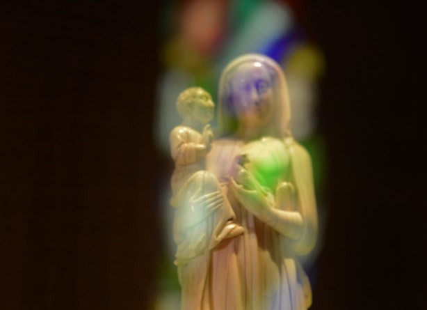 a small white statue of Mary holding the baby Jesus, lit by light coming through a stained glass window behind it 