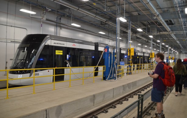 inside the new maintance building, a new grey, black and white crosstown train on display, people walking past it and taking pictures