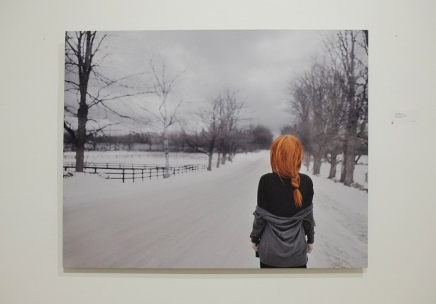 a large photo of a red headed woman, long hair, in a braid, standing on a snow covered road with her back to the camera, in the countryside