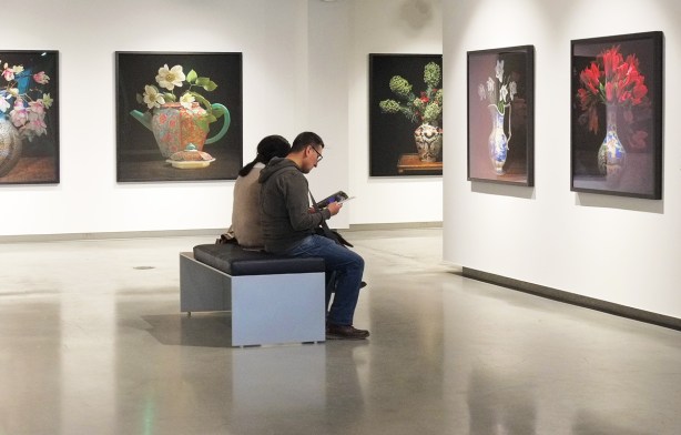 people sitting on a bench in a gallery, reading, large photos of flower bouquets on the walls around them 
