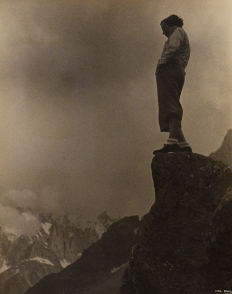 anold photo by Ilse Bing of a woman standing on a rock high upon a mountian. She's looking down over the valley below