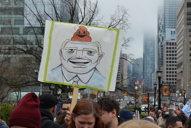 a person holds up a sign that is a drawing of Doug Ford's head with a poop emoji on top of his head 