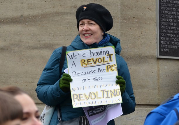 a woman in a blue coat and black beret holds a sign that says We're having a revolt because the PC's are so revolting 