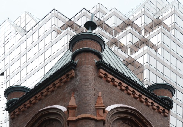 the top corner of an old red brick building with a green roof, with a new glass building behind it 