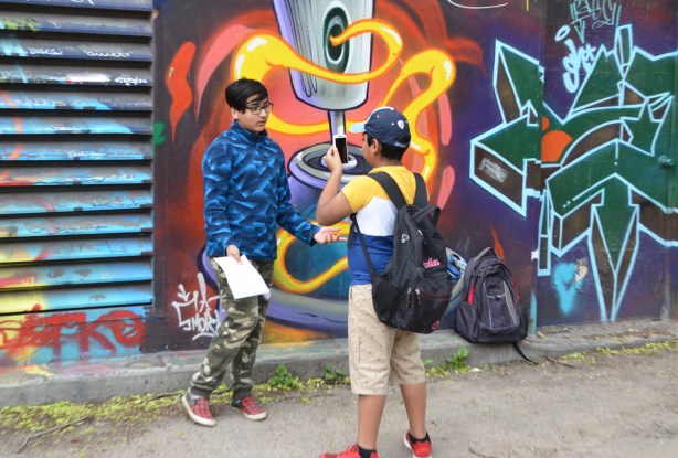 a boy in shorts and a baseball cap is filming another boy in blue fleece and camo pants as he is talking in front of a mural in graffiti alley
