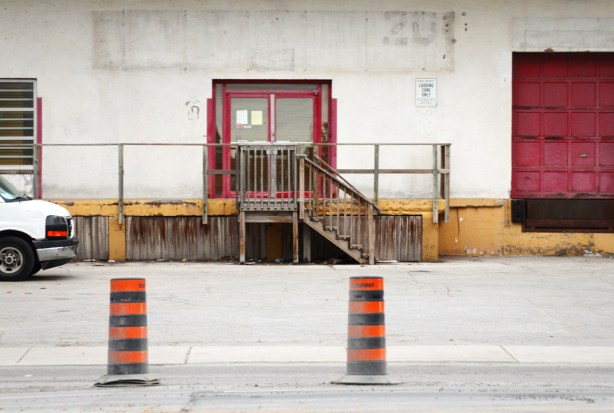 two orange and black traffic contruction cones on the street in front of an old white industrial building with red door frames and a wood loading dock with yellow trim