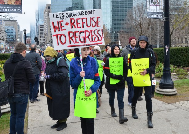 a group of artists with bright yellow and light green signs walks as a group towards a protest in front of Queens Park, the woman at the front also has a sign on a stick that says We refuse to regress