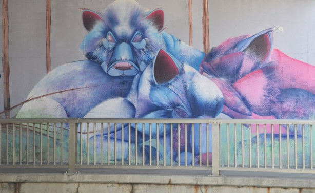 part of a mural by Christopher Ross on an underpass wall in pinks and blues, a large blue bear