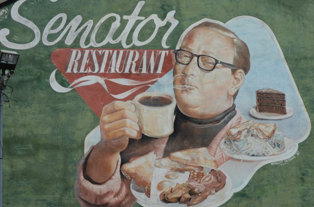 large mural on the side of the Senator restaurant, a man in glasses holds a steaming cup of coffee with plates of food in front of him, by his shoulder, bacon and eggs with toast as well as a plate with a sandwich and a side of salad. A third plate has a piece of chocolate cake 