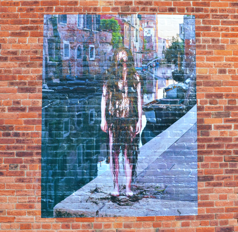 large photo on a brick wall, taken by a canal in Venice, of a woman covered from head to toe in black weeds as she stands beside the canal