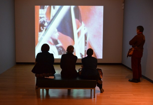 4 people watching a video on a large screen, one person is standing while 3 people are sitting on a bench with their backs to the camera