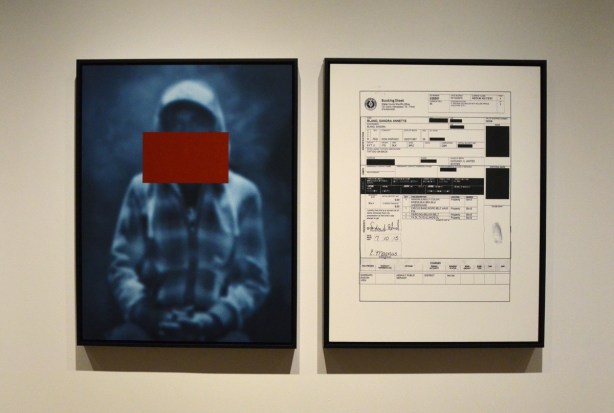 park of an exhibit in a gallery showing the picture of a black boy with a large red rectangle acros his face, beside it is an enlargement of the arrest record of a black man in Ferguson Missouri 