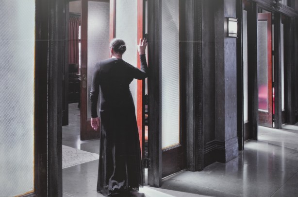 large photo on a wall outside, of a woman in a long black dress, back to camera, one hand on door sill as she stands in open doorway, by Carrie Mae Weems 