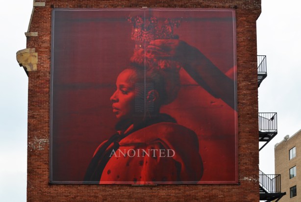 a large red photo of a woman being crowned, sitting in profile, the word anointed is written in large letters on the picture. Mounted on the side of a red brick building