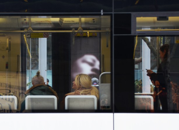 people sitting in a streetcar with their back to the window, can see large photo on exhibit on opposite sidewalk through the windows of the streetcar 