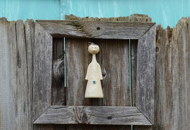 a stylized figurine, round head on conical body, screwed onto a wood fence and surrounded by a wood frame 