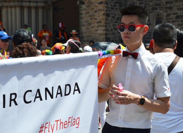 a young asian man in white button down shirt, black pants, black bowtie, and red sunglasses, stands beside an air canada banner, getting ready for pride parade 