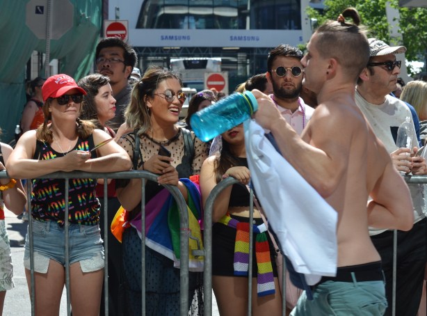 women behind metal barricades at pride parade, as a topless man walks past 