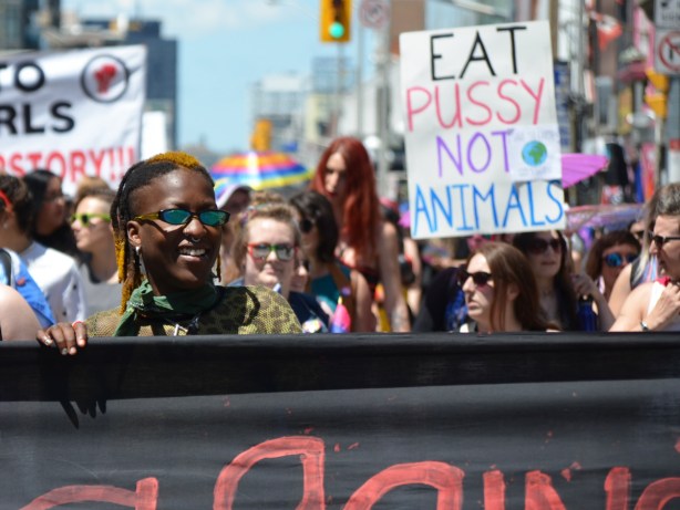 black woman holding on to a banner in a parade with crowd behind her, someone holding a sign that says eat pussy not animals