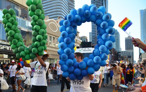 two young men in Pride Parade, one has a giant O made of blue balloons and the other is carrying a giant U made of green umbrellas 