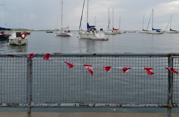 sailboats moored out in Lake Ontario, in the foreground is a metal railing with a string of Canadian flags tied to it 