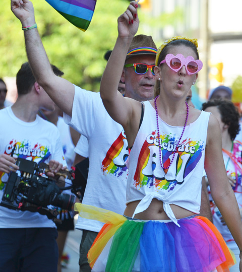 a couple in the pride parade, each wearing white t shirts that say celebrate all, and each with one arm in the air, woman has pink sunglasses and man has rainbow sunglasses and fedora 