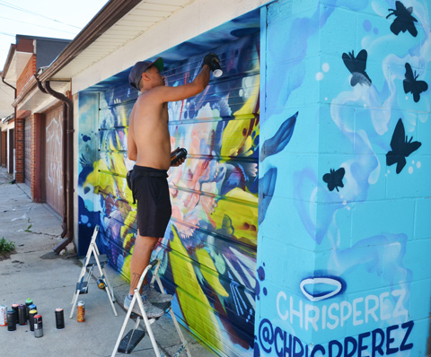 a man on a short ladder spray painting a mural on a garage door in an alley, Chris Perez