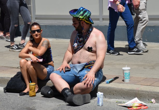 a couple sits on a sidewalk watching the dyke march, he is wearing a blue and green hat and has tape X's on his nipples