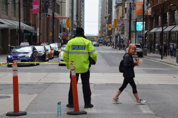a police man in a bright yellow jacket stands in front an orange cone and yellow police tape to block off Yonge Street. He's directing traffic, to make cars turn on Queen street. A woman with orange hair is walking across the street , just went in front of the police man
