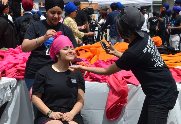 a young person in a black T-shirt is directing a woman sitting to get the best pose for a photo, woman is having a pink turban wrapped around her head by another woman at Turbanup event at Yonge Dundas square