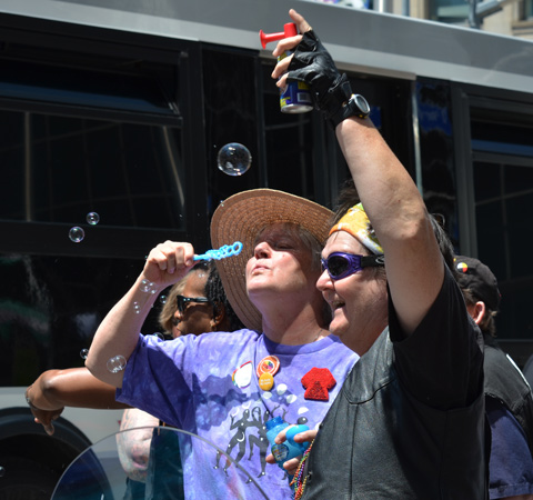 two women, one with brown large rimmed hat blowing bubbles and the other with arm raised and canned horn noise maker in her hand