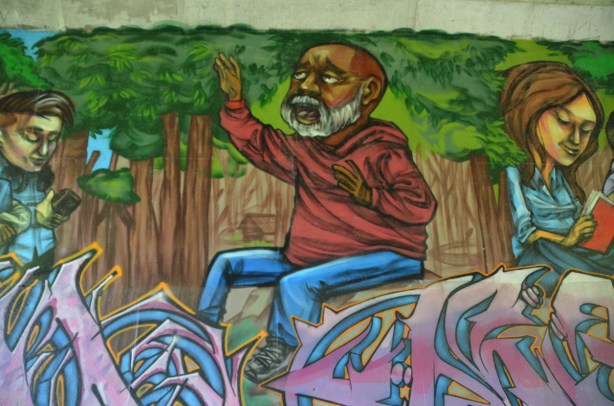 part of an elicser mural beside West Highland Creek, under Lawrence Ave., an older man in a red shirt with a white beard and moustache, beside him is a girl reading (in the mural)