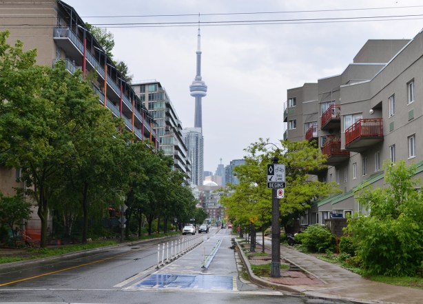 looking east along Queens Quay West from the very western end of the ctreet towards the CN Tower and downtown. Lowrise residential units on either side of the street, bike lane, small trees, wet, raining 