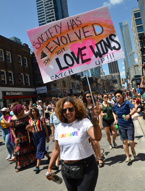 women walkin in dyke march down Yonge Street, one woman holds a sign that says Society evolved, love wins, catch up!