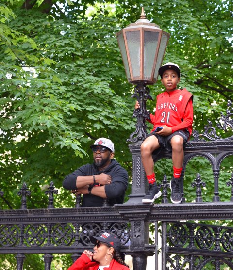 Raptors championship parade day, , a young boy sits up high on the Osgoode Hall fence, beside a light, his father is close by 