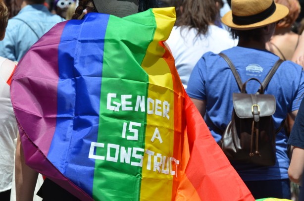 person with rainbow flag draped on their back, white tape has been used to make the words gender is a construct on the flag