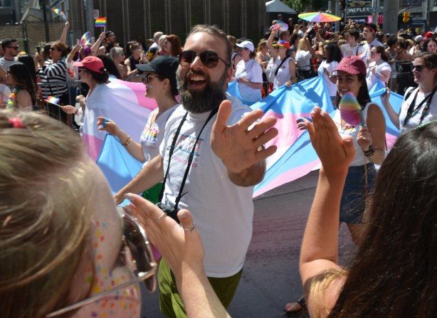 a man with a beard and moustache, and camera around his neck, walking in pride parade, reaches out his hand, hands of spectators reaching out to him 