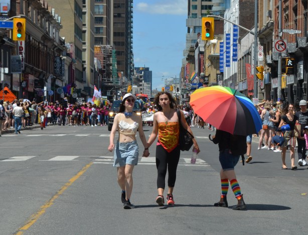 a person under a very large rainbow umbrella is beside two women who are walking while holding hands, last section of dyke march parade is behind them