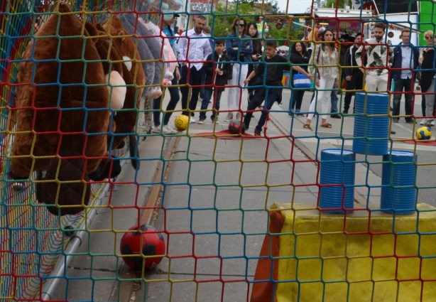 behind a net, a line of people watches as a boy starts to kick a soccer ball towards three blue cans, game to try to knock the cans over 