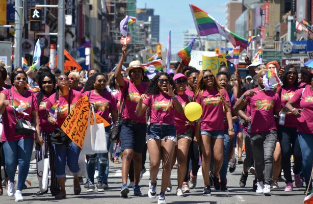 a group of women in maroon coloured t shirts are walking together in dyke march, singing and cheering, with signs, flags and yellow balloon
