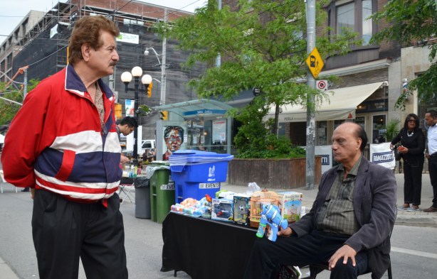 a man seated by a table on the sidewalk, starts to shoot a man in a red jacket with a bubble blowing gun shaped like a blue fish 