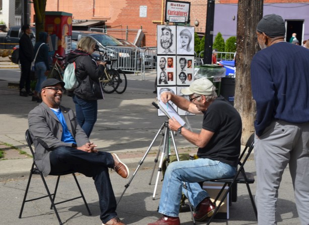 a man is starting to draw a caricature of a black man who is seated, laughing, on a chair. Another black man watches 