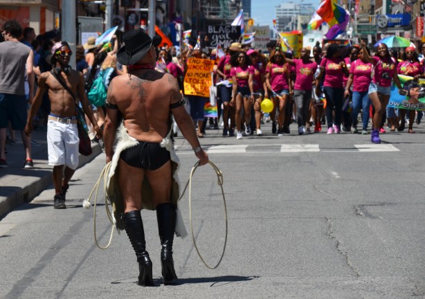man with whip, high black boots, and bare back, stands with back to camera and facing a group of women in the dyke march
