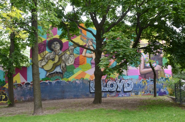 large mural on the side of a building beside green space, with a couple of trees in front 